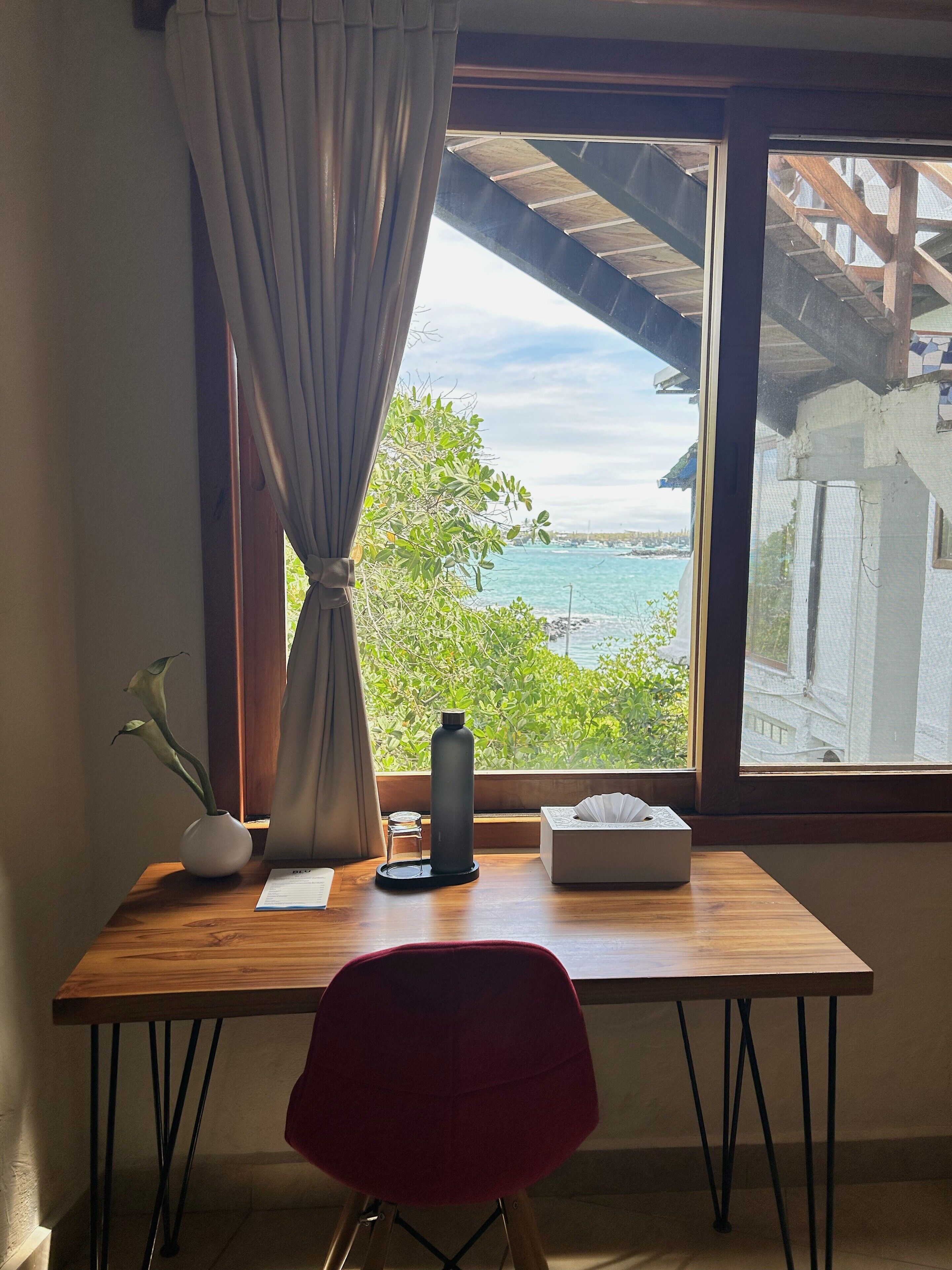 Una habitación con un escritorio de madera, una silla roja, un ventanal con vista al mar, una planta en maceta y una caja de pañuelos.