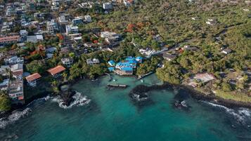 An aerial view of a coastal town with a prominent blue-roofed building surrounded by lush greenery and a clear turquoise sea.