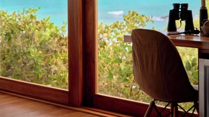 A room with a large window overlooking a turquoise sea and palm trees. There is a wooden desk with binoculars and a chair.