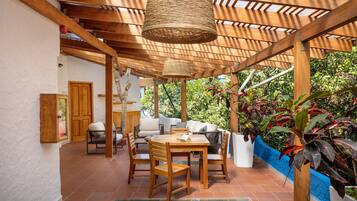 A covered patio area with a wooden table and chairs, a sofa, and a hanging light fixture.