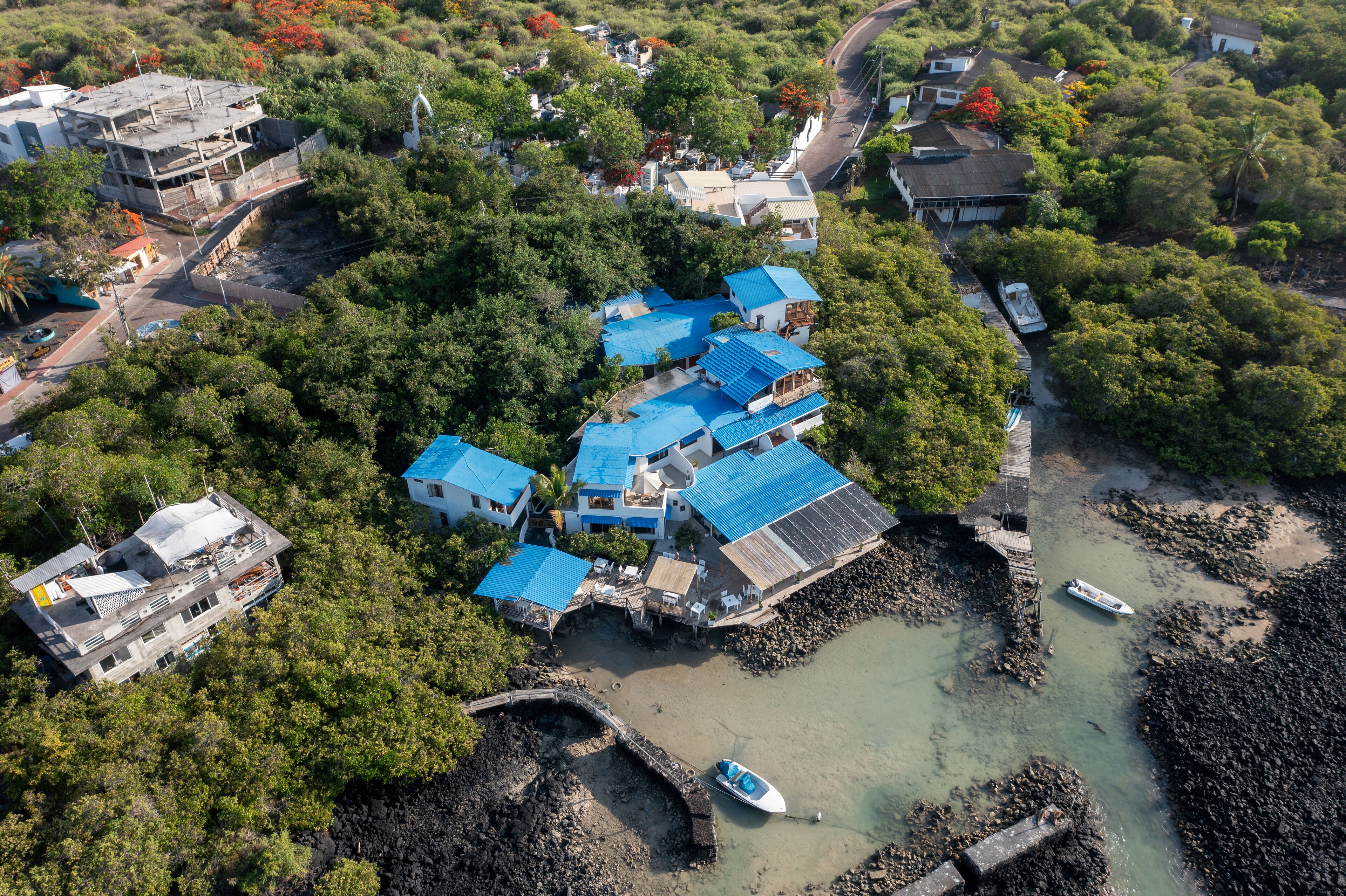 An aerial view of a coastal area with multiple buildings, including a prominent structure with blue roofs, surrounded by lush greenery and a rocky shoreline with boats.