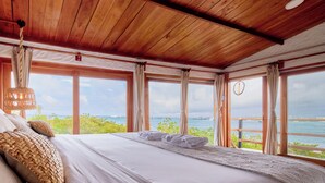 A bedroom with a wooden ceiling, large windows, a bed with white linens, and a view of the sea.