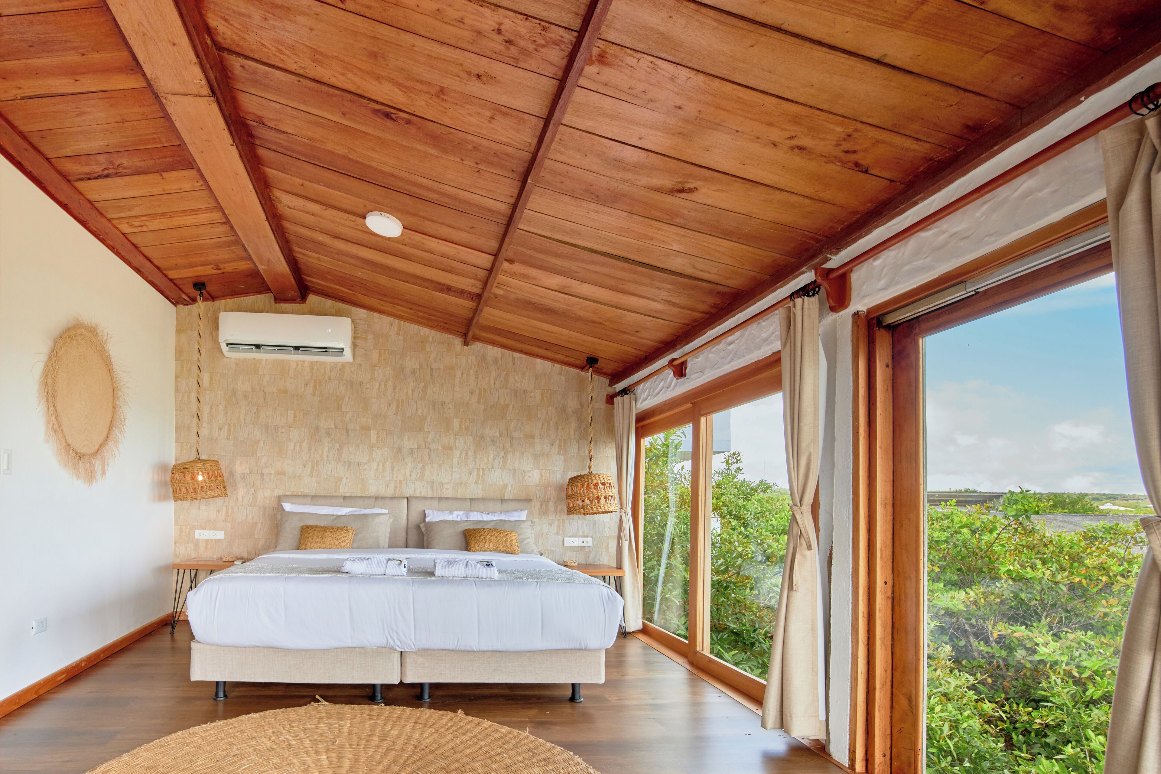 A bedroom with a large bed, wooden ceiling, and a view of greenery through large windows.