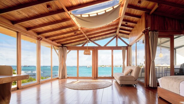 A spacious room with a large window overlooking the sea, wooden flooring, a round rug, a bed, and a seating area.