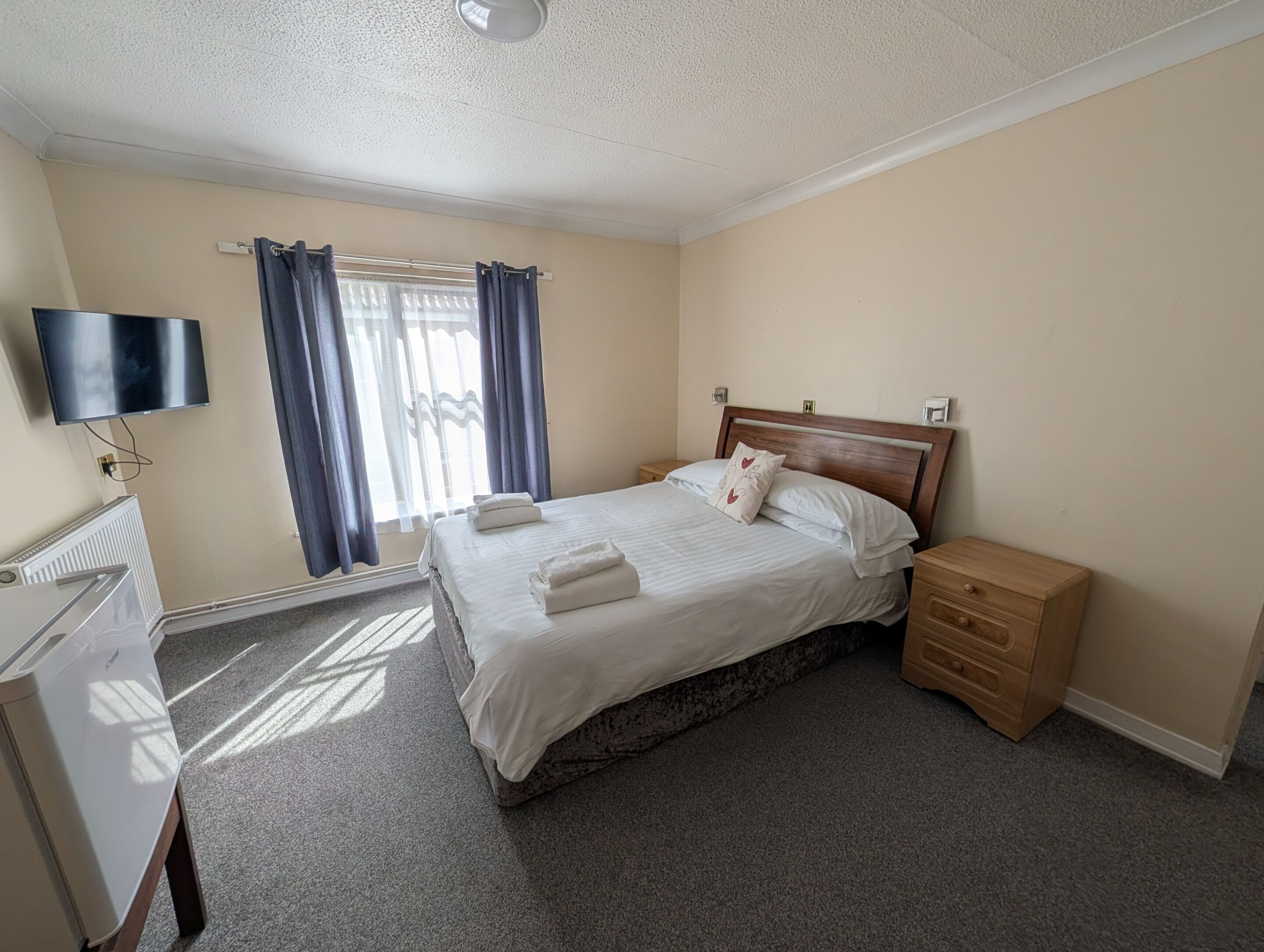 Habitación de hotel con cama, mesita de noche, televisor de pantalla plana montado en la pared, radiador y ventana con cortinas.