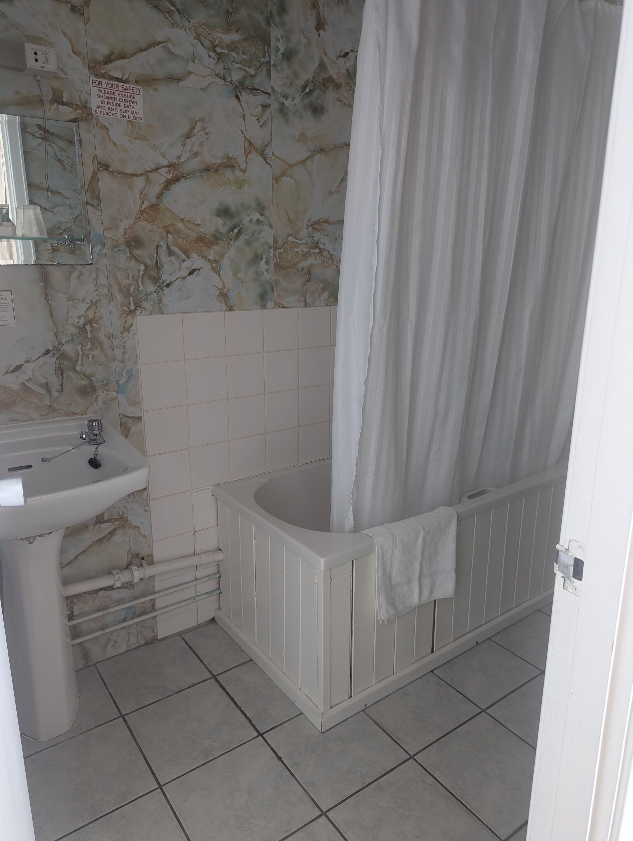 Un baño con pared de mármol, una bañera con cortina, un lavamanos y una toalla blanca en el radiador.