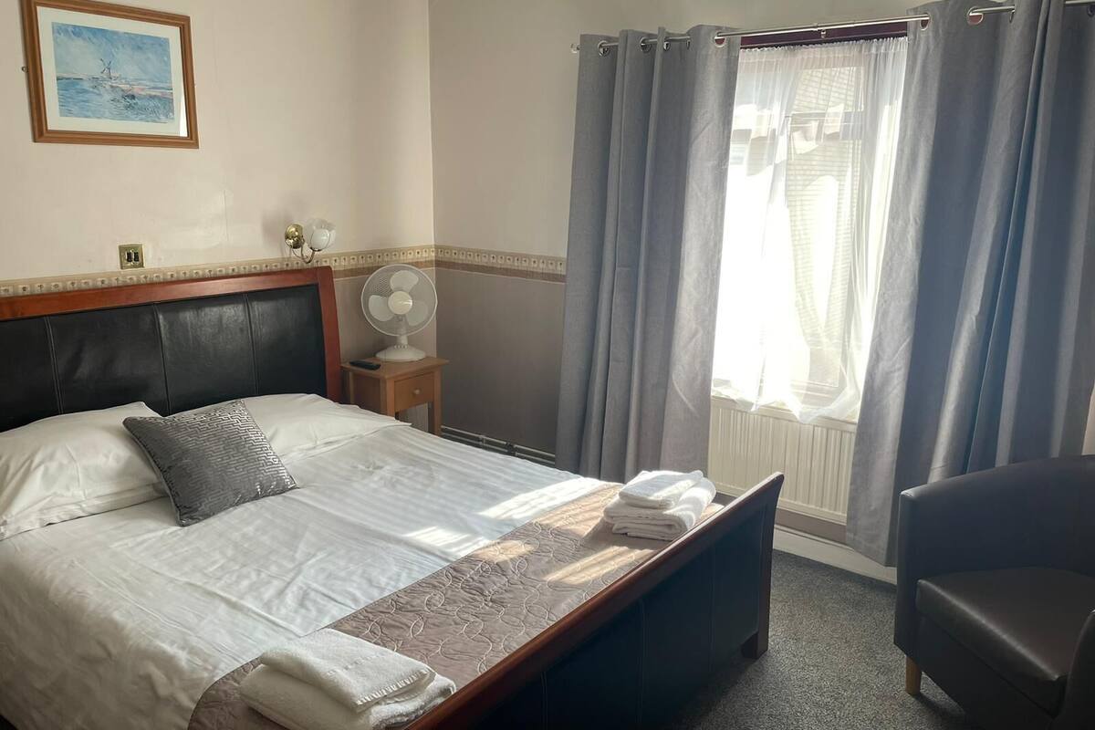 A hotel room with a bed, a bedside table with a fan, a chair, and a window with curtains.