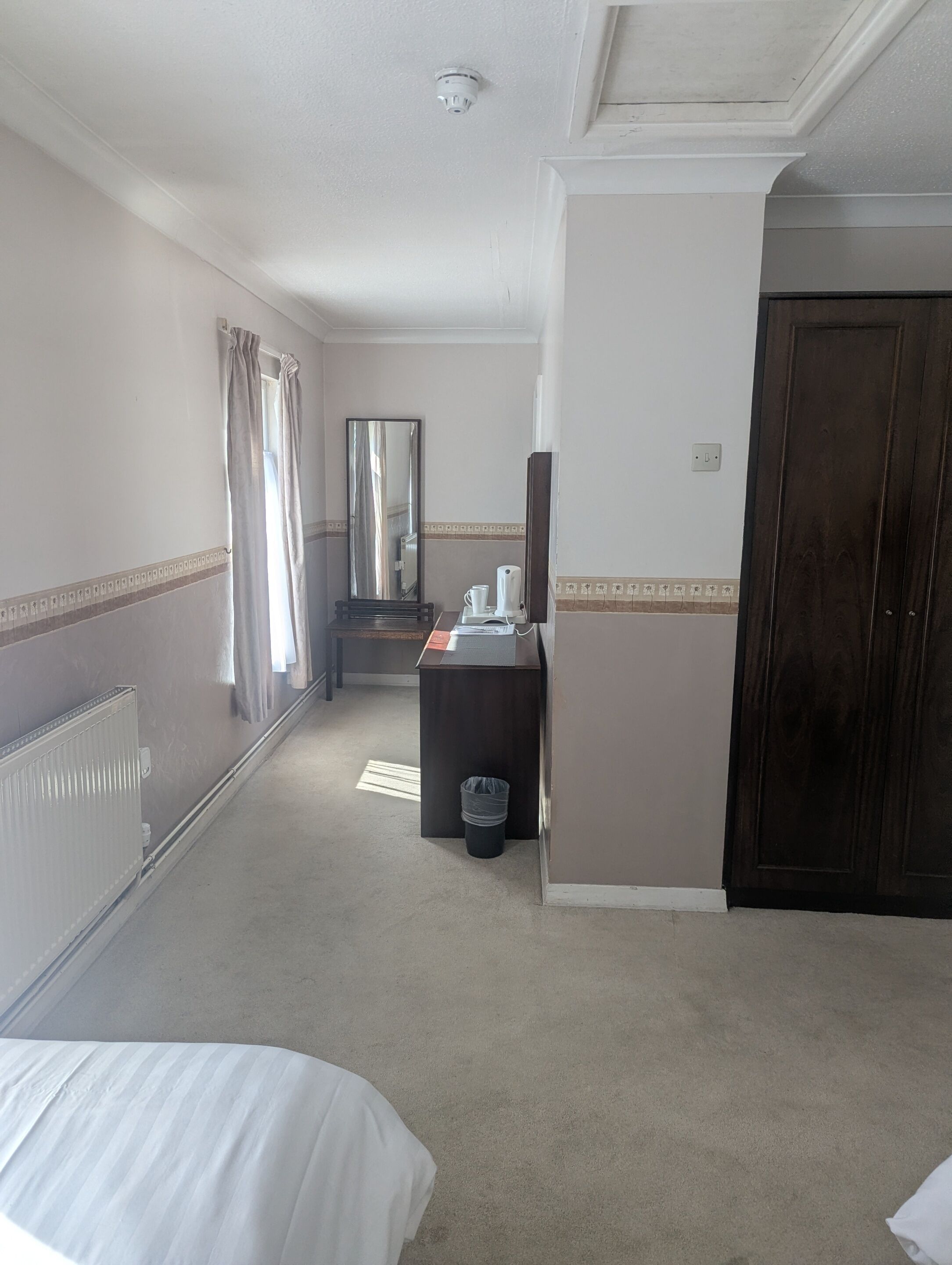 A hotel room with a bed, a desk with a mirror, a radiator, a waste bin, and a door.