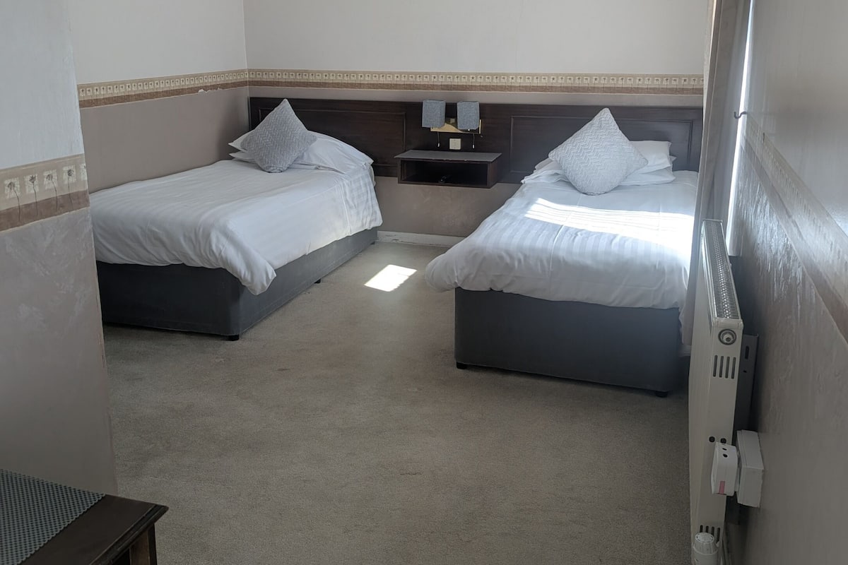 A hotel room with two beds, a small bedside table, a radiator, and a power outlet.