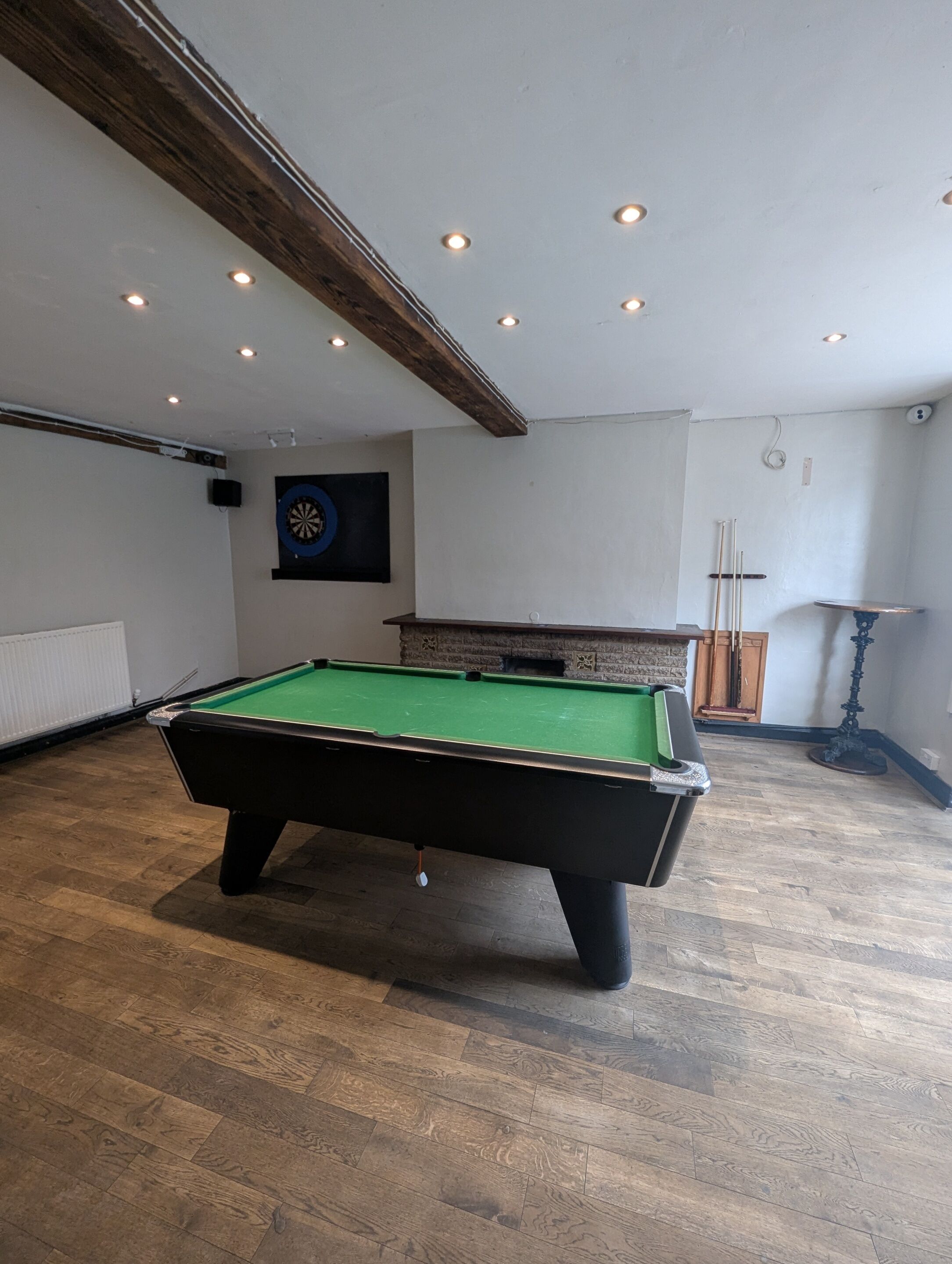 A pool table in a room with wooden flooring, recessed ceiling lights, a dartboard, and a small table with a candle.