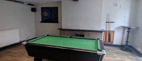 A pool table in a room with wooden flooring, recessed ceiling lights, a dartboard, and a small table with a candle.