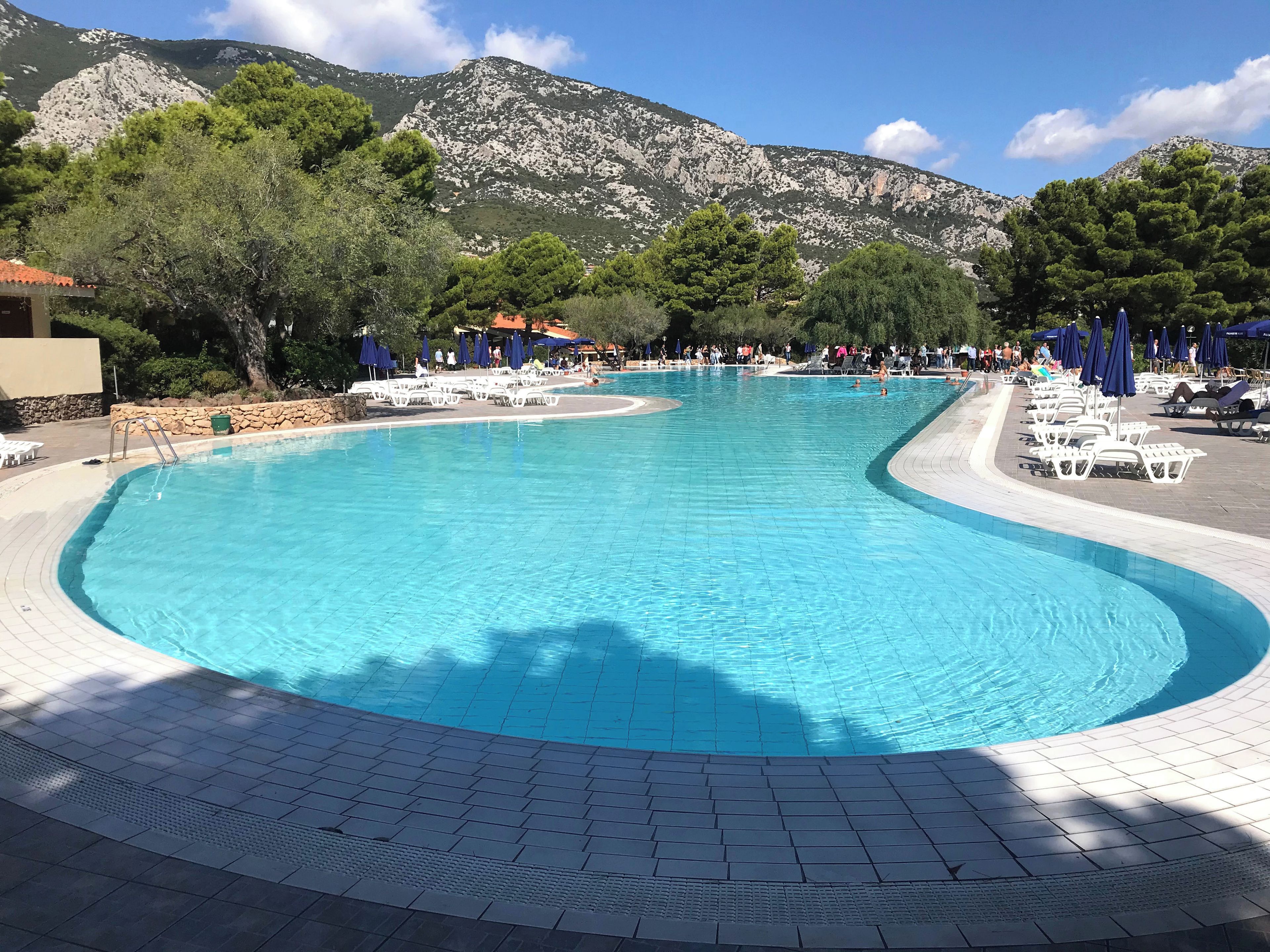 Ein Pool mit gewölbter Kante, umgeben von Liegestühlen und Sonnenschirmen, mit Bergen im Hintergrund.