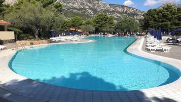Ein Pool mit gewölbter Kante, umgeben von Liegestühlen und Sonnenschirmen, mit Bergen im Hintergrund.