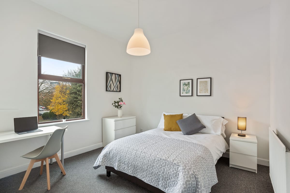 Quarto com cama, escrivaninha, cadeira, mesa de cabeceira e janela com vista para árvores.