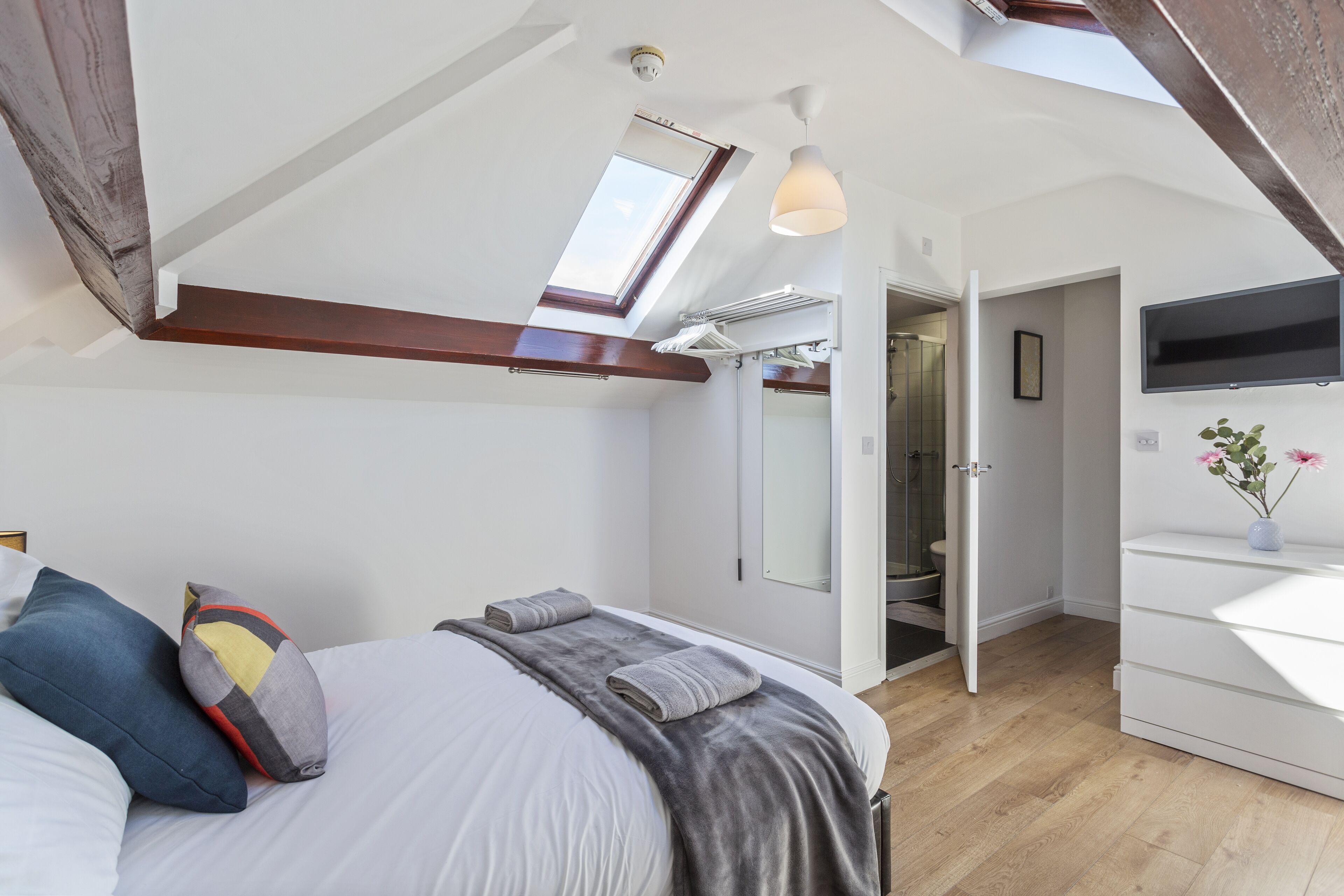A bedroom with a skylight, a bed with pillows, a bedside table, and a TV on the wall.