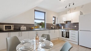 A modern kitchen with a dining area, featuring a dining table set for four, a microwave, a toaster, and a large window providing natural light. - River Rooms Nottingham (Nottingham)