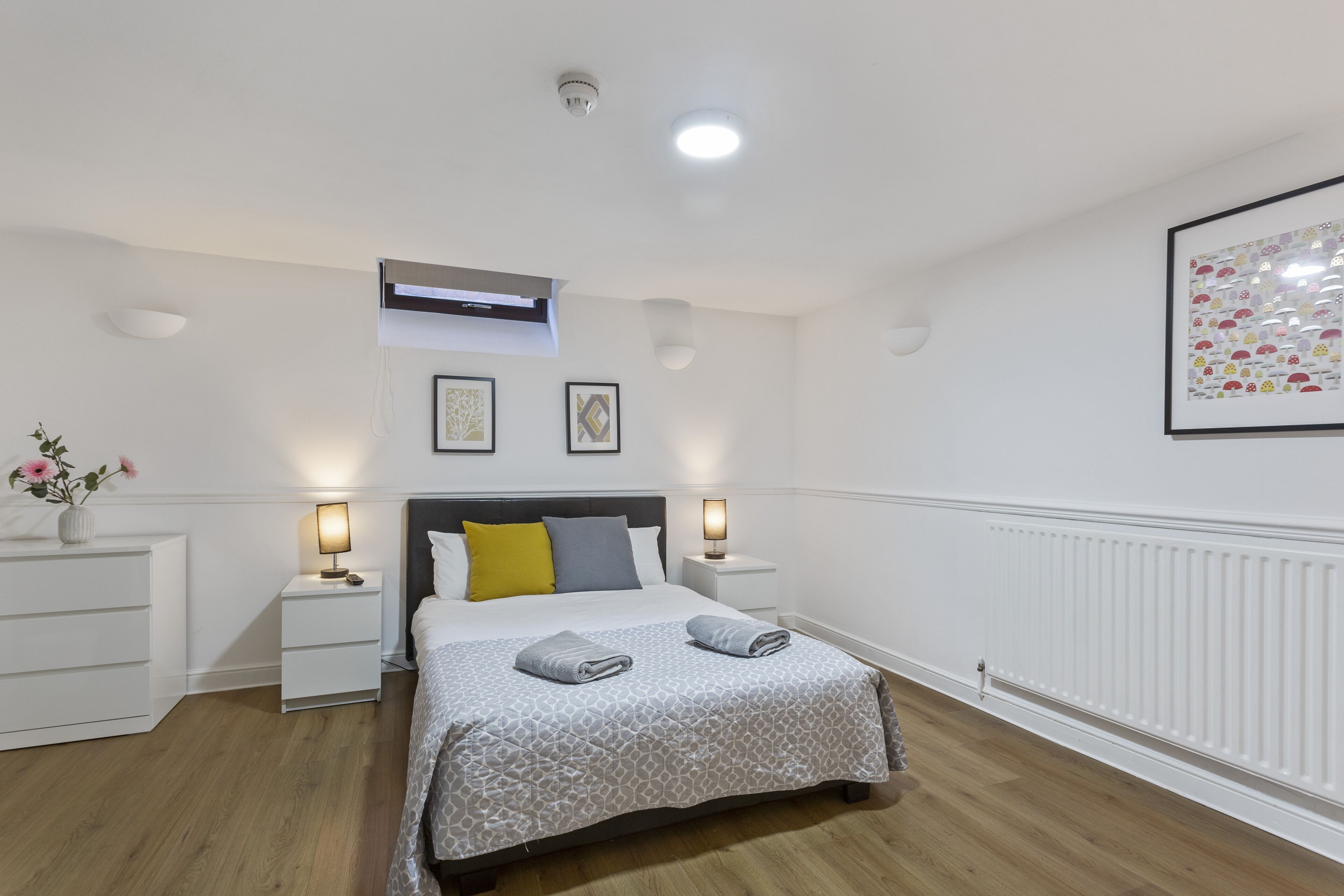 A bedroom with a bed, bedside tables, a nightstand, a wall-mounted air conditioner, a framed picture, and a radiator.
