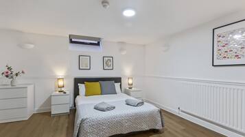 A bedroom with a bed, bedside tables, a nightstand, a wall-mounted air conditioner, a framed picture, and a radiator.