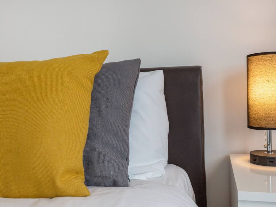 A bed with three pillows in yellow, gray, and white, and a bedside table with a lamp.
