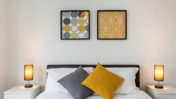 Een net opgemaakt bed met twee kussens, een witte muur met twee kunstwerken in lijsten, en twee bedlampen op witte nachtkastjes.