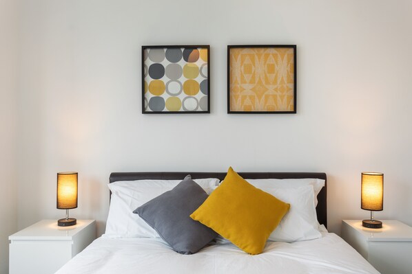 Una cama bien hecha con dos almohadas, una pared blanca con dos obras de arte enmarcadas y dos lámparas de noche sobre mesitas blancas.