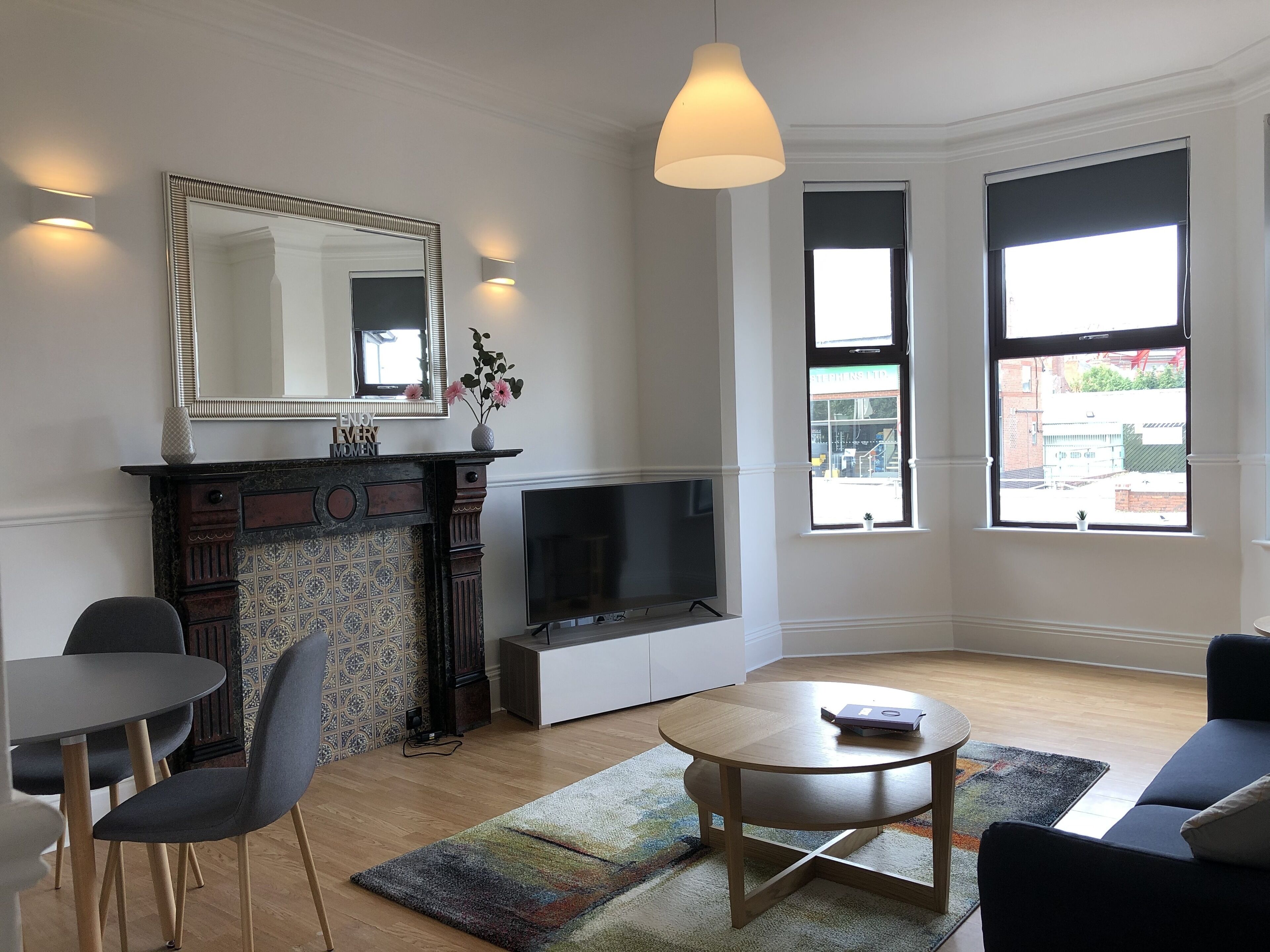 Een moderne woonkamer met een open haard, een flatscreen televisie, een eethoek met stoelen en grote ramen met uitzicht op de straat.