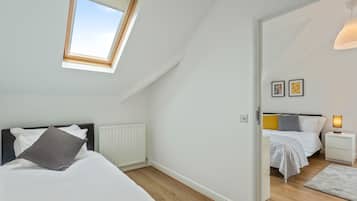 Una camera da letto con un lucernario, un letto con biancheria bianca, un comodino con una lampada e vista su un'altra stanza attraverso una porta aperta.