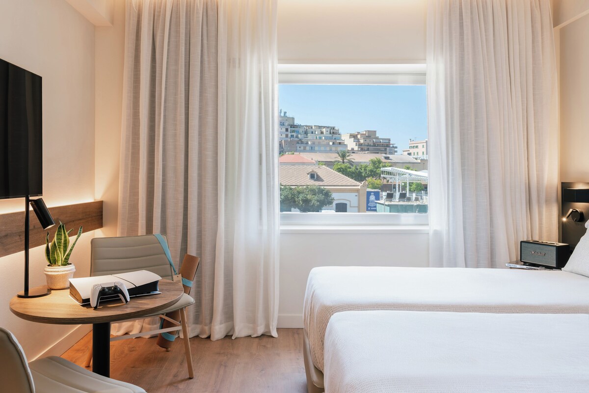 Une chambre d’hôtel avec un lit, un bureau et une chaise, une petite table avec une plante, et une vue sur la ville par la fenêtre.