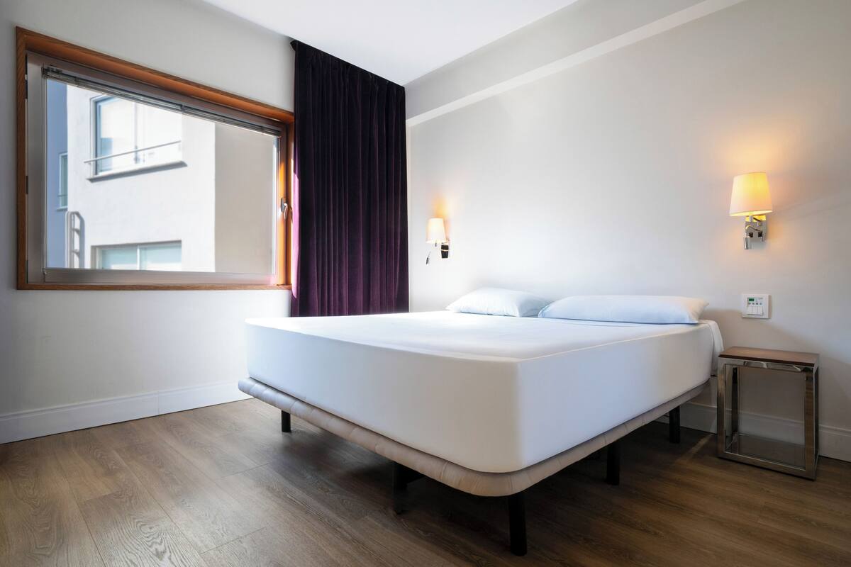 Una habitación de hotel moderna con una cama grande, una ventana con cortinas, una lámpara de noche y una mesita.