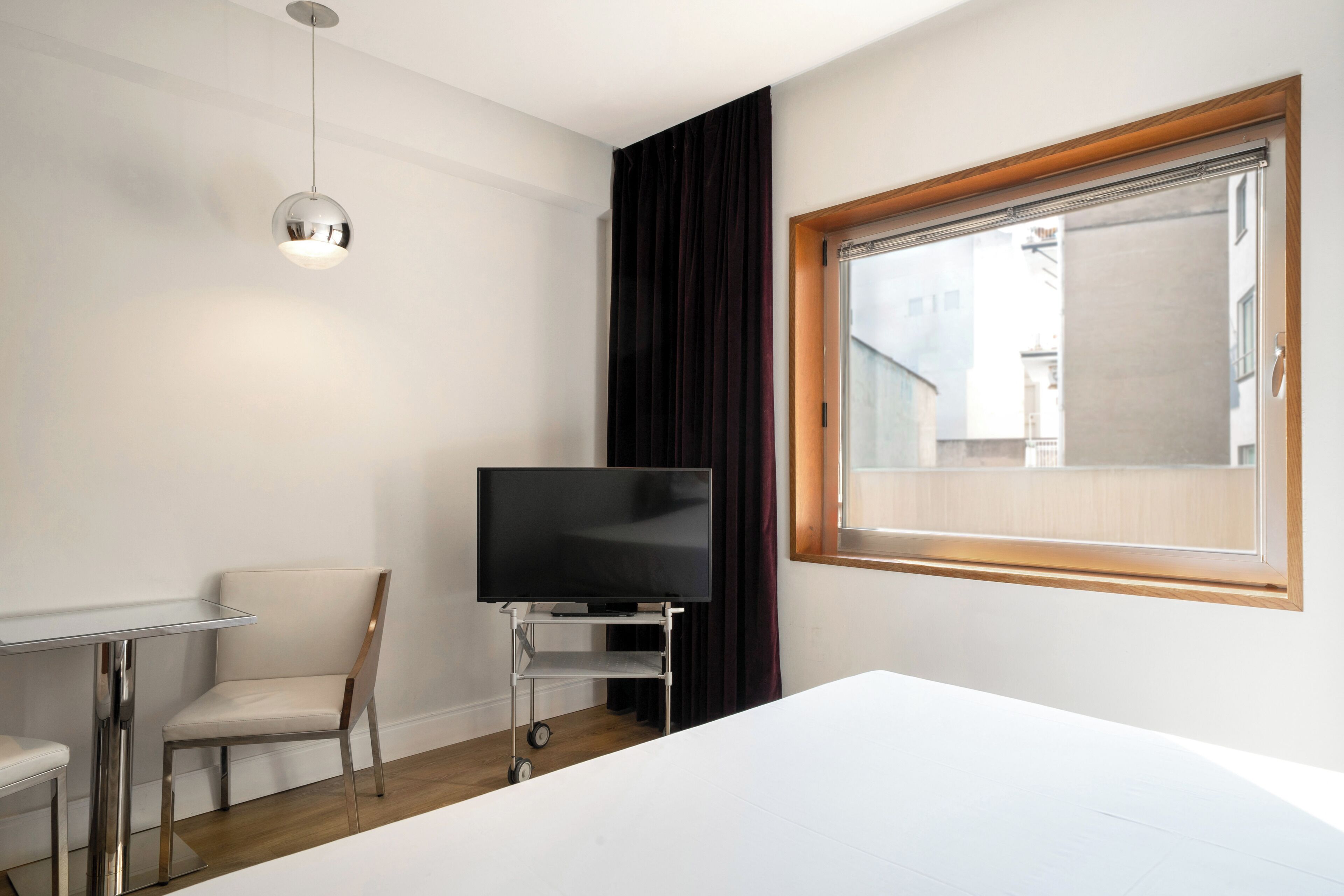 Habitació d'hotel amb un llit, un televisor sobre un prestatge, una cadira, una taula petita i una finestra amb cortines.