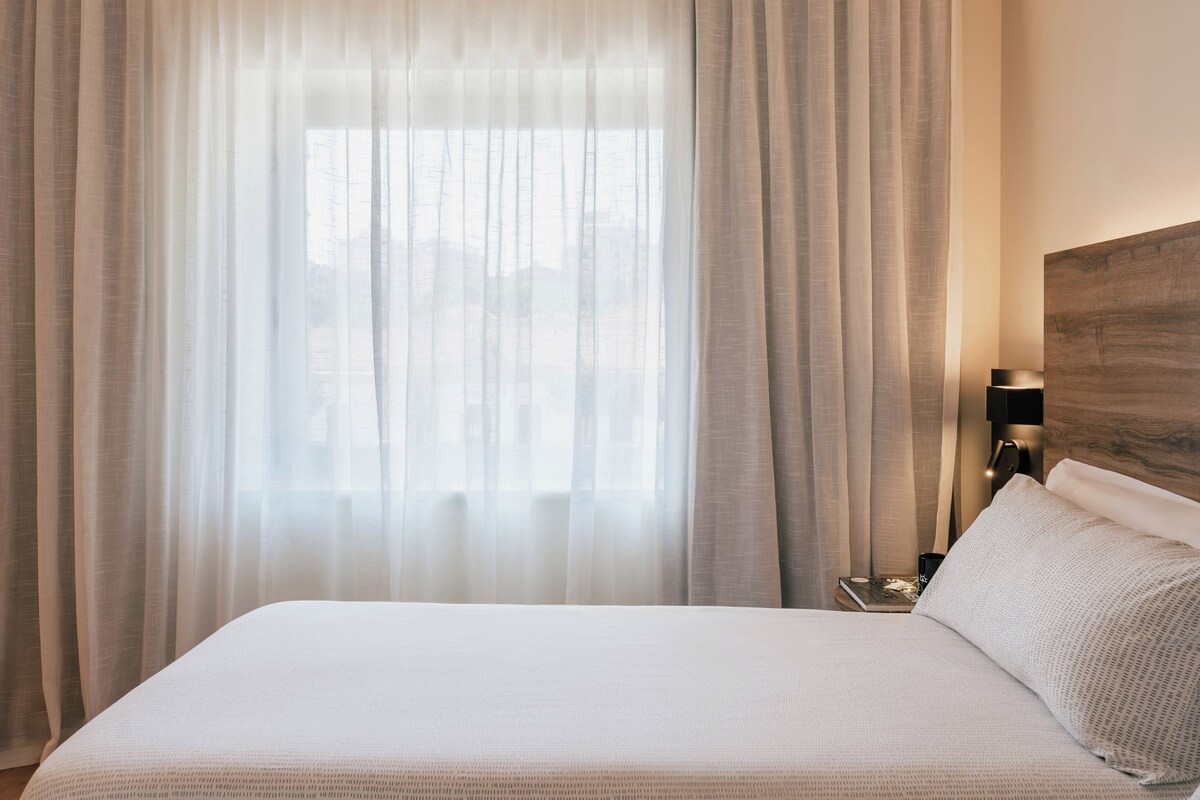 Une chambre d’hôtel avec un lit, des rideaux, une table de chevet et une lampe fixée au mur.