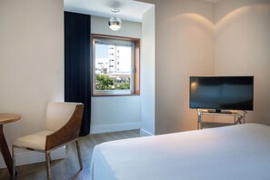 Une chambre d’hôtel avec un lit, un téléviseur posé sur un meuble, une chaise et une fenêtre donnant sur des immeubles.