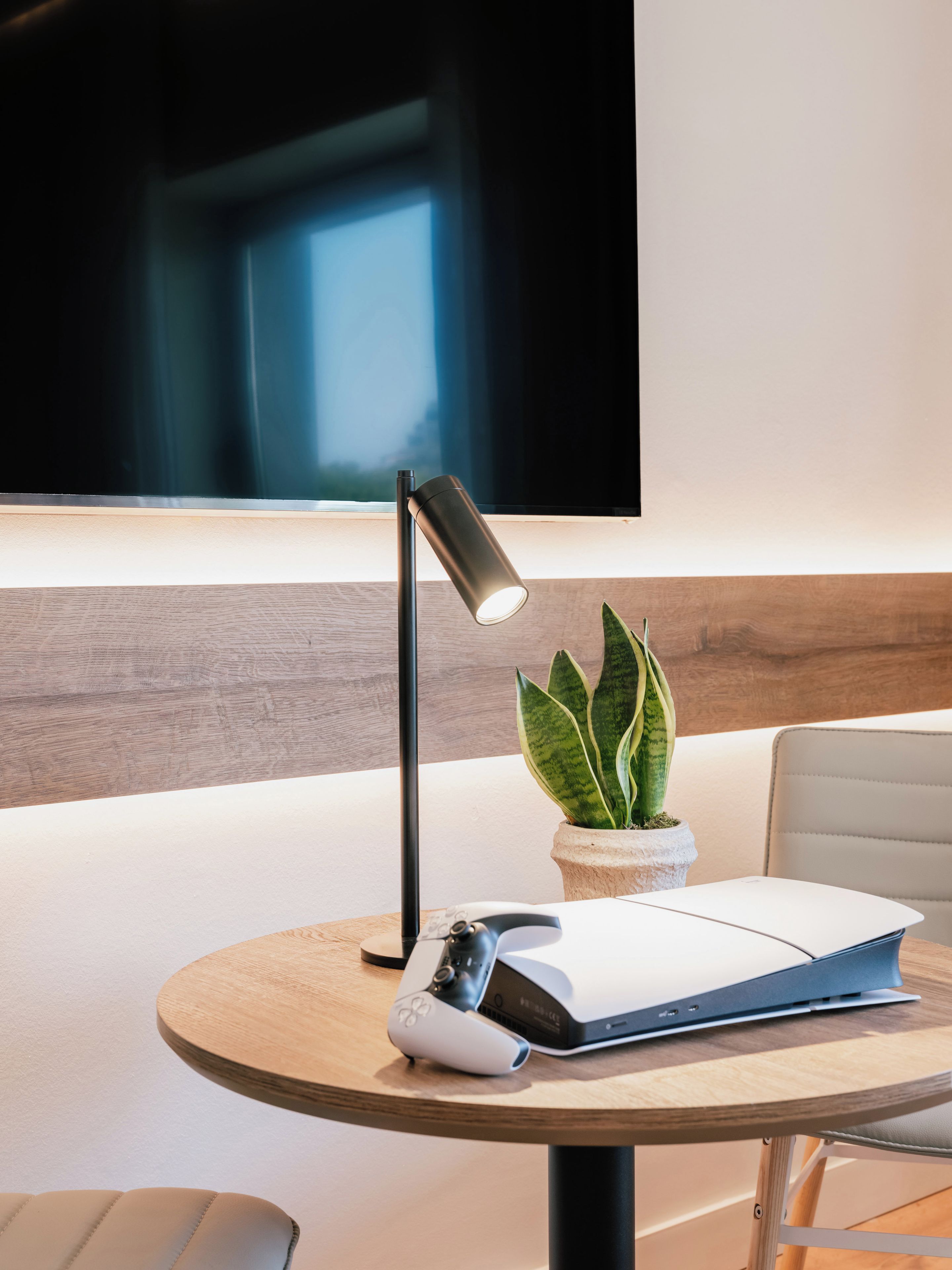 Una habitación moderna con una mesa redonda, una consola de videojuegos, una planta y un televisor de pantalla plana montado en la pared.