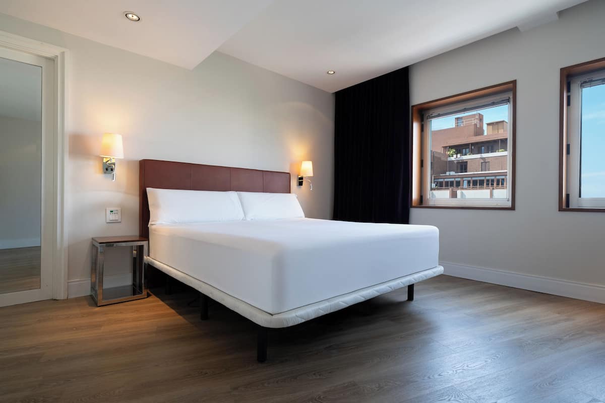 Una habitación de hotel moderna con una cama grande, dos lámparas de noche, un ventanal con vistas a edificios y suelo de madera.