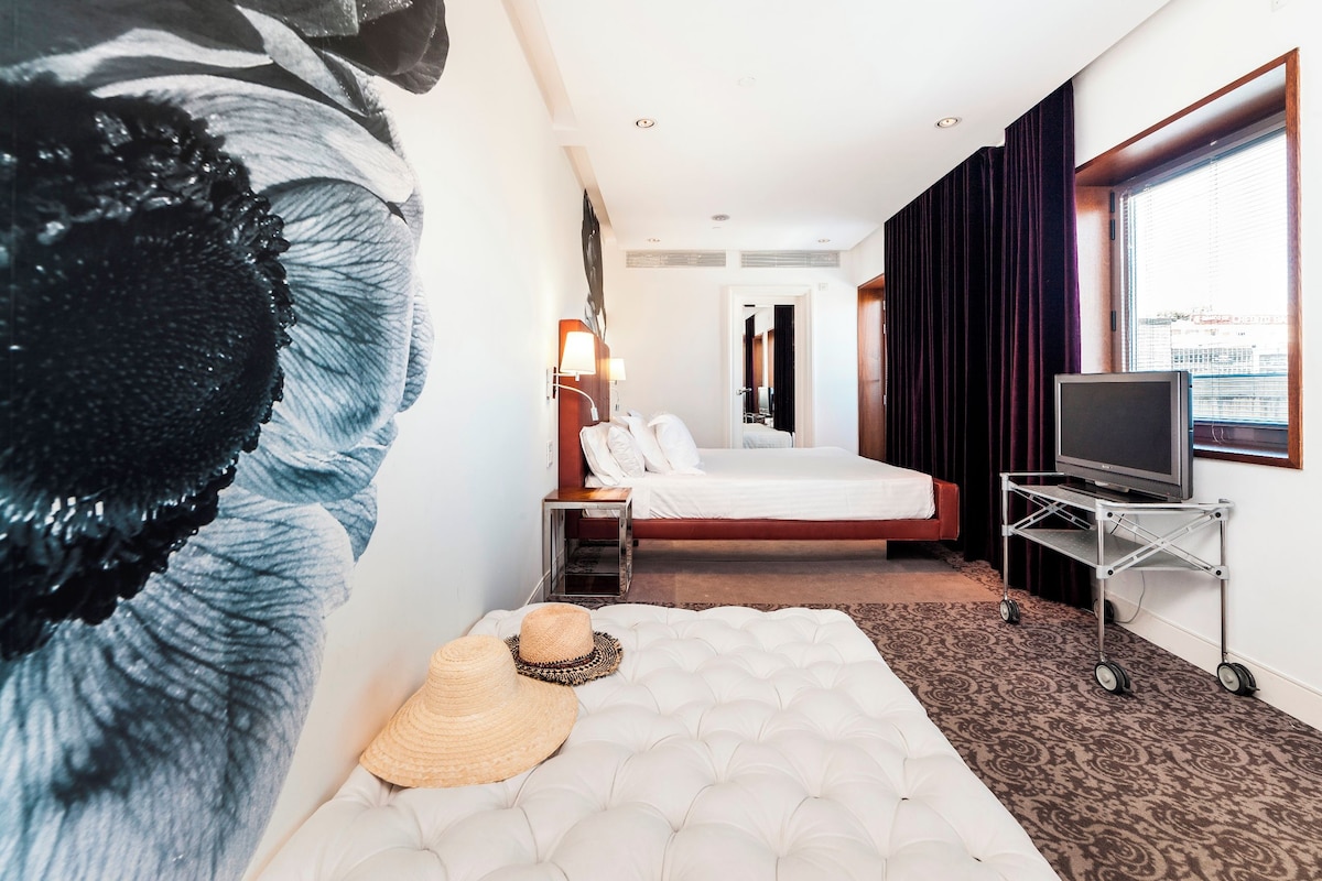 Una habitación de hotel con una cama grande, un televisor sobre un mueble bajo, una mesita de noche, una lámpara de pared y un cuadro de gran tamaño.