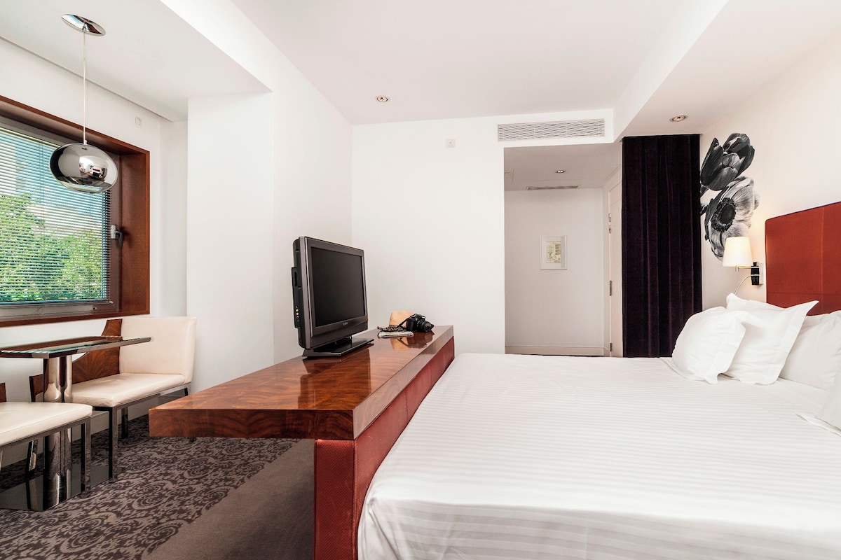Una habitación de hotel con cama, escritorio con televisor, una silla y ventana con persianas.