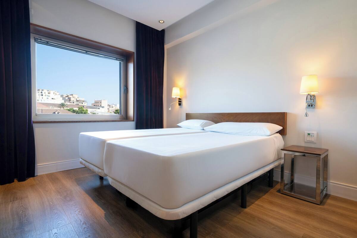 Habitación de hotel con una cama grande, dos mesitas de noche y un ventanal con cortinas.