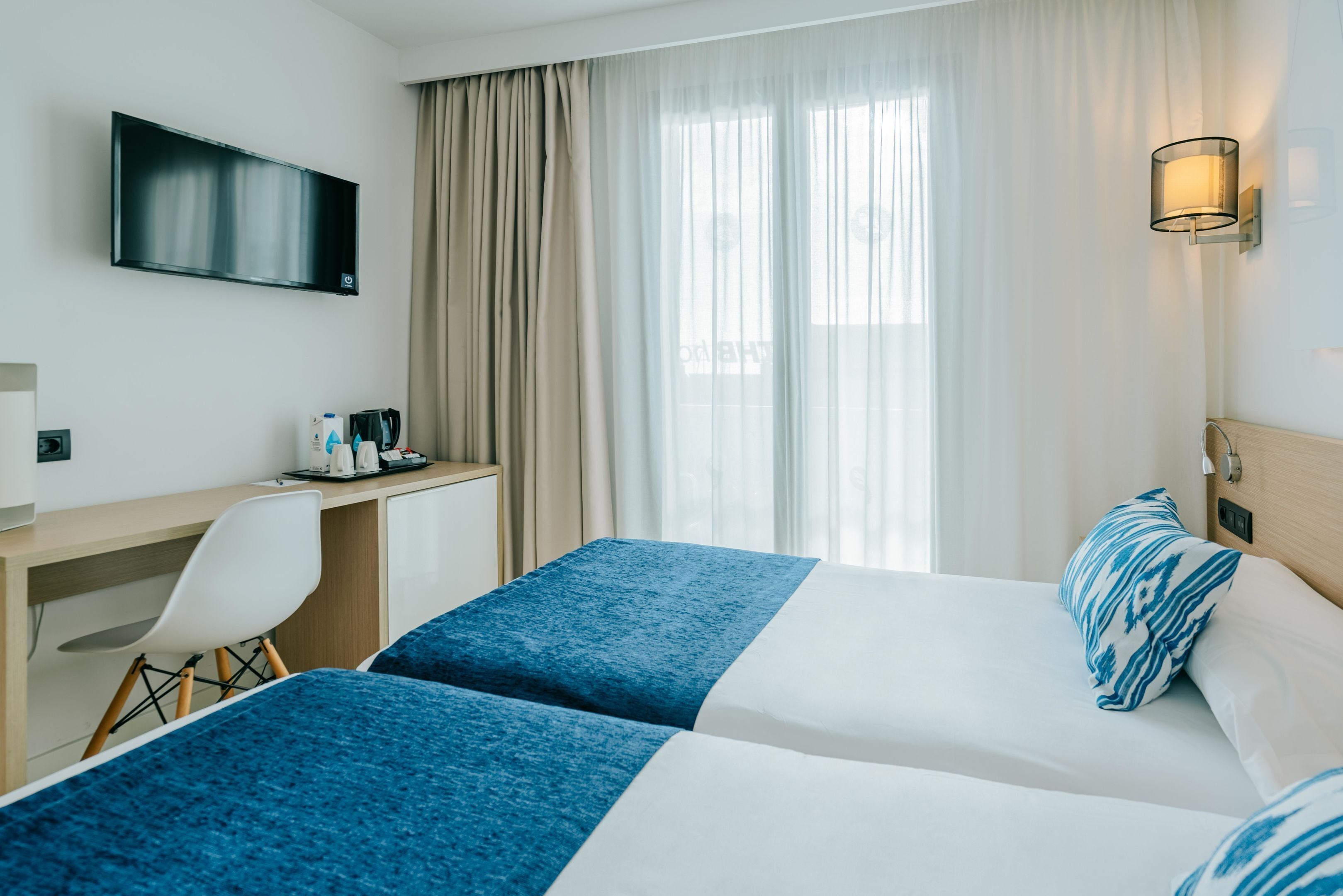 A hotel room with two beds, a desk, a chair, a TV, and a window with curtains.