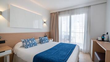 A hotel room with a large bed, a desk, and a chair. There is a window with curtains, a wall-mounted light, and a flat-screen TV.