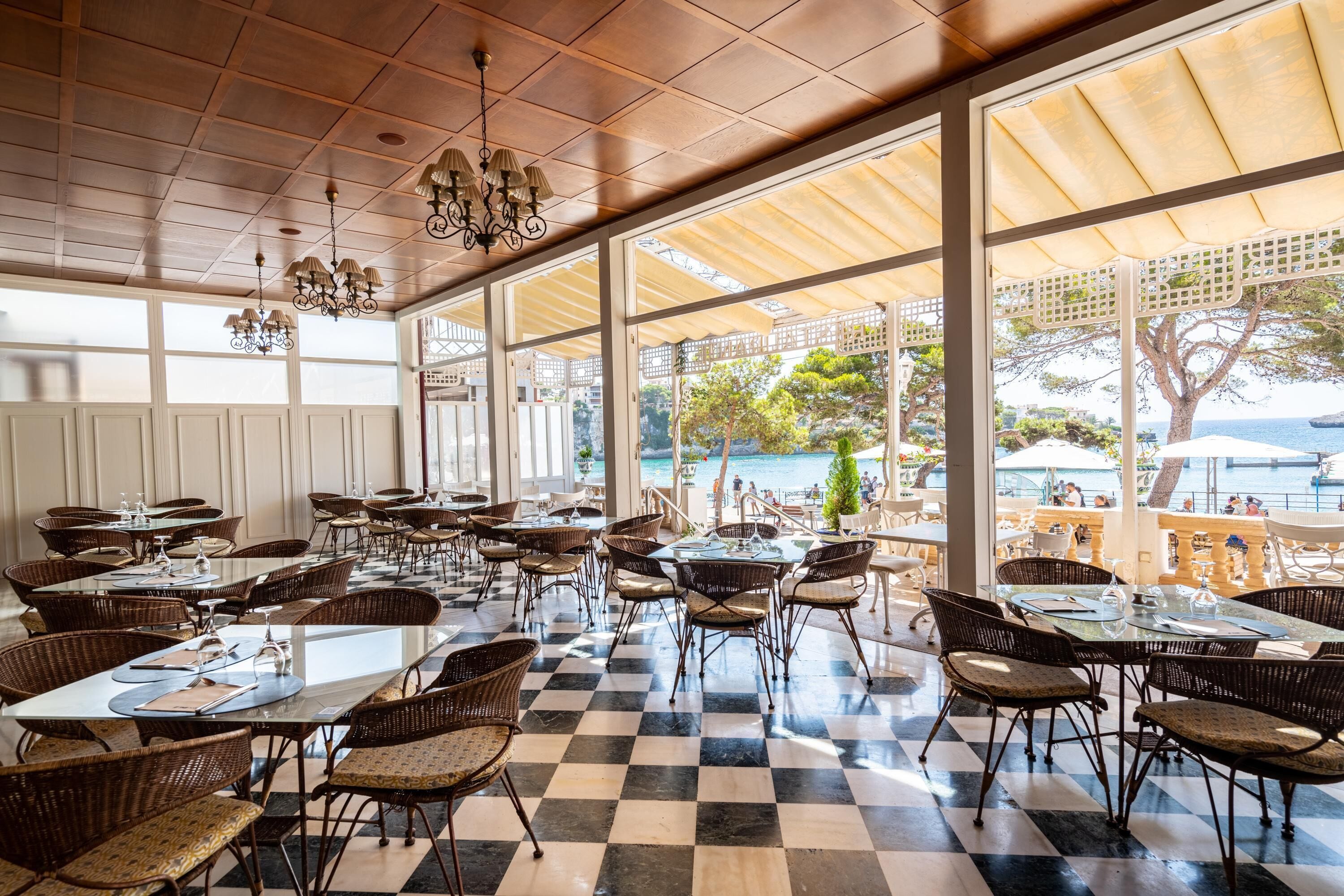 Un amplio comedor interior con suelo a cuadros, sillas de mimbre y mesas dispuestas para comer. Ventanas grandes ofrecen vistas al mar y una zona de estar al aire libre.