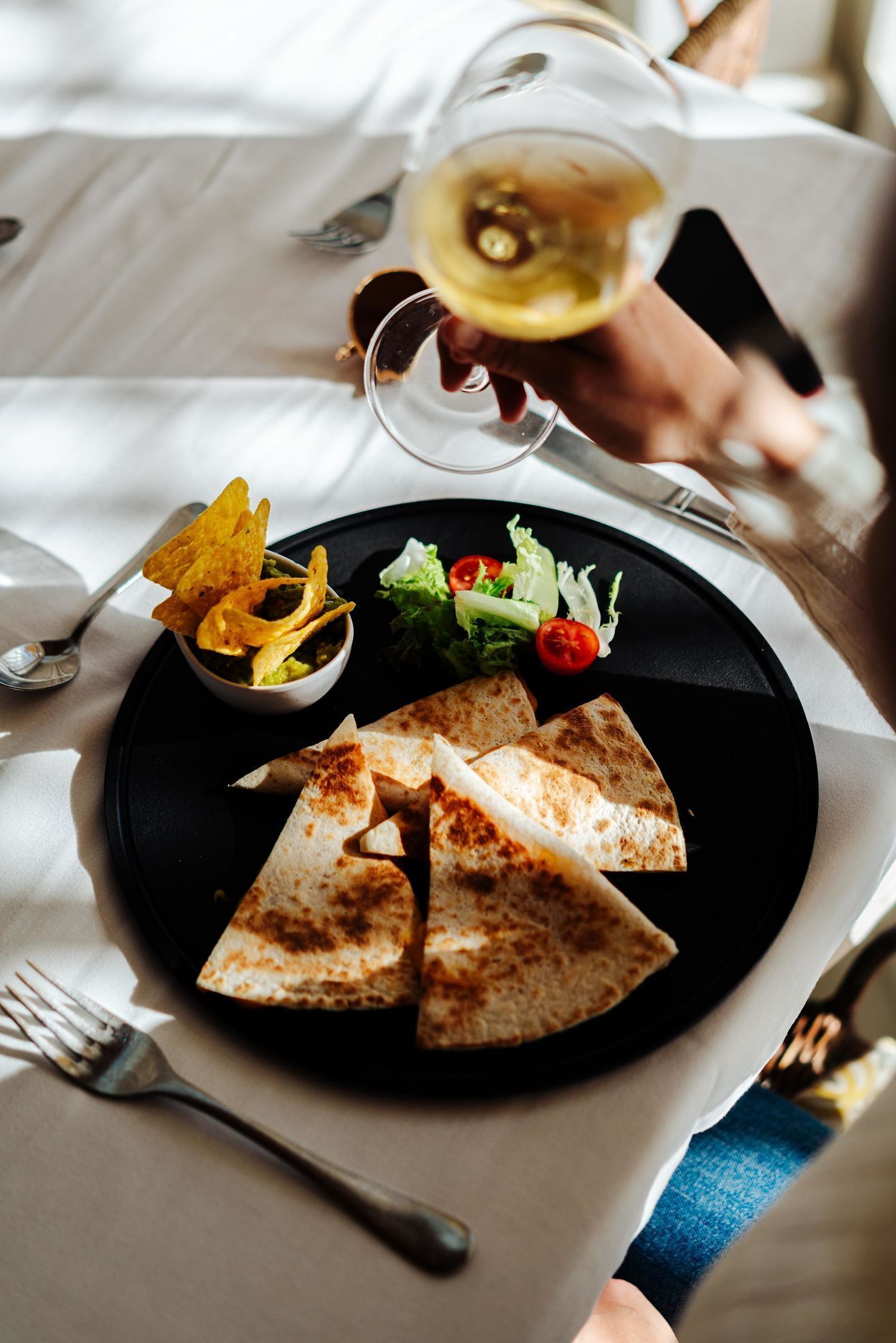 Un plato negro con tres quesadillas, un tazón pequeño de guacamole y una ensalada sobre un mantel blanco.