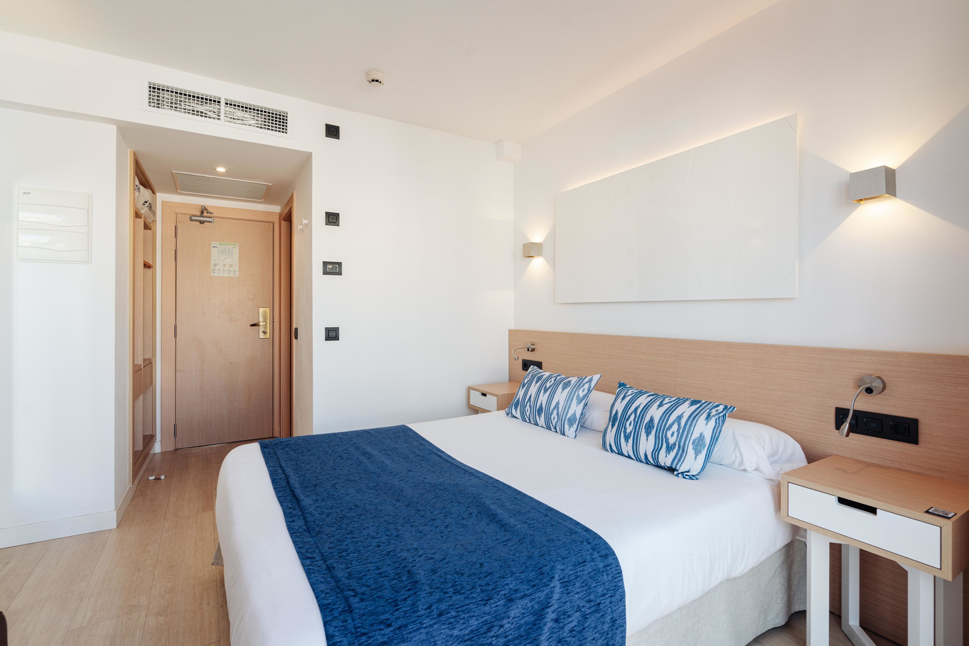 A hotel room with a bed, bedside table, and a wall-mounted light fixture.