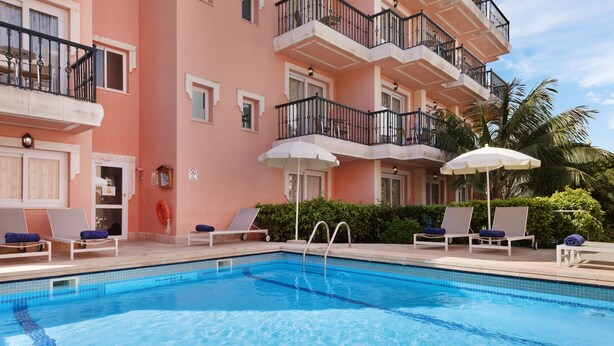 Piscina com cadeiras de praia e guarda-sóis, rodeada por um edifício rosa com varandas.