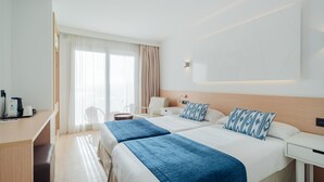 Quarto de hotel com duas camas, uma escrivaninha, uma cadeira, um abajur e uma janela com cortinas.