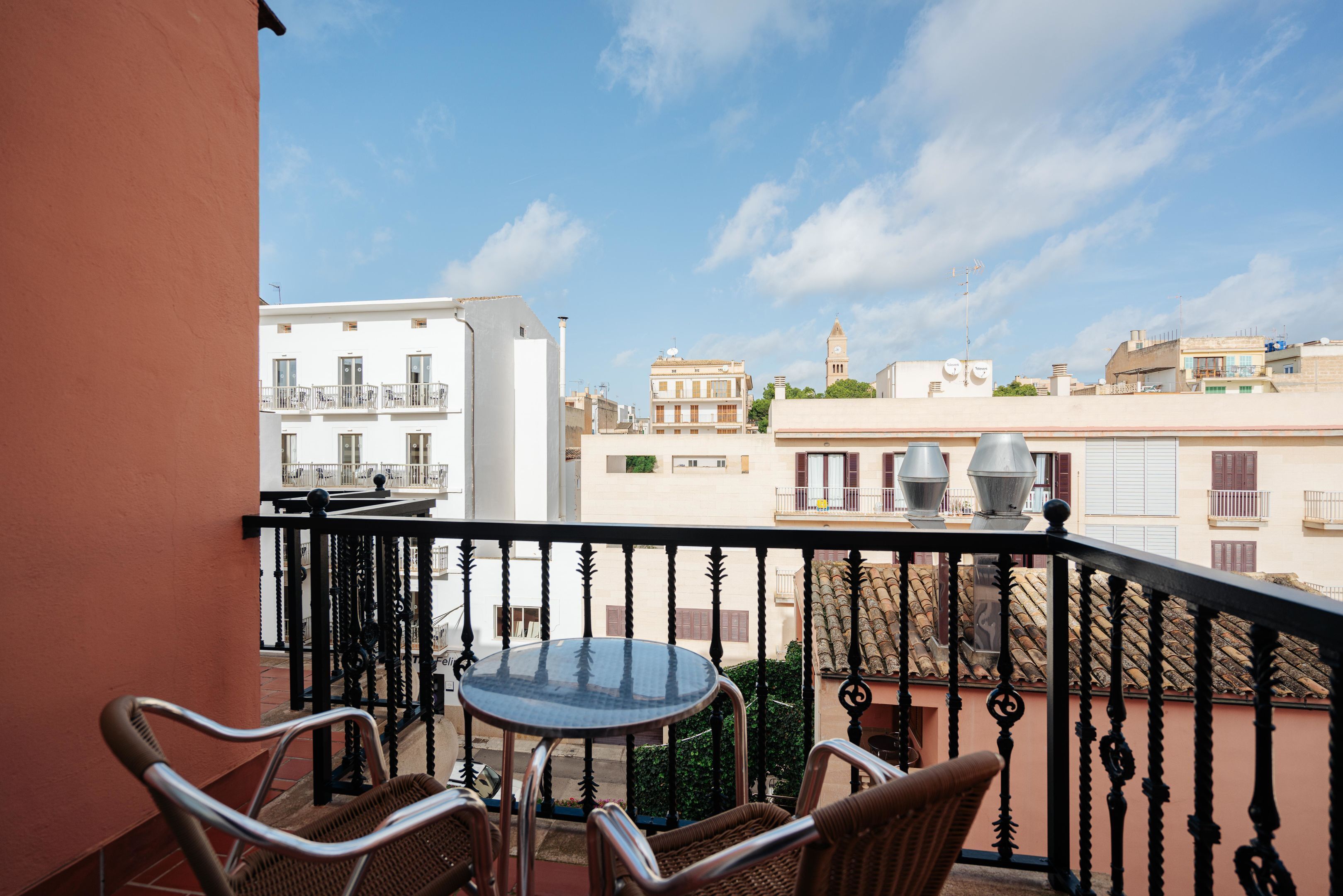 Een balkon met rieten stoelen, een glazen tafel en uitzicht op omliggende gebouwen.