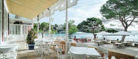 Un café al aire libre con mesas y sillas blancas, una zona de estar cubierta y vista al mar.