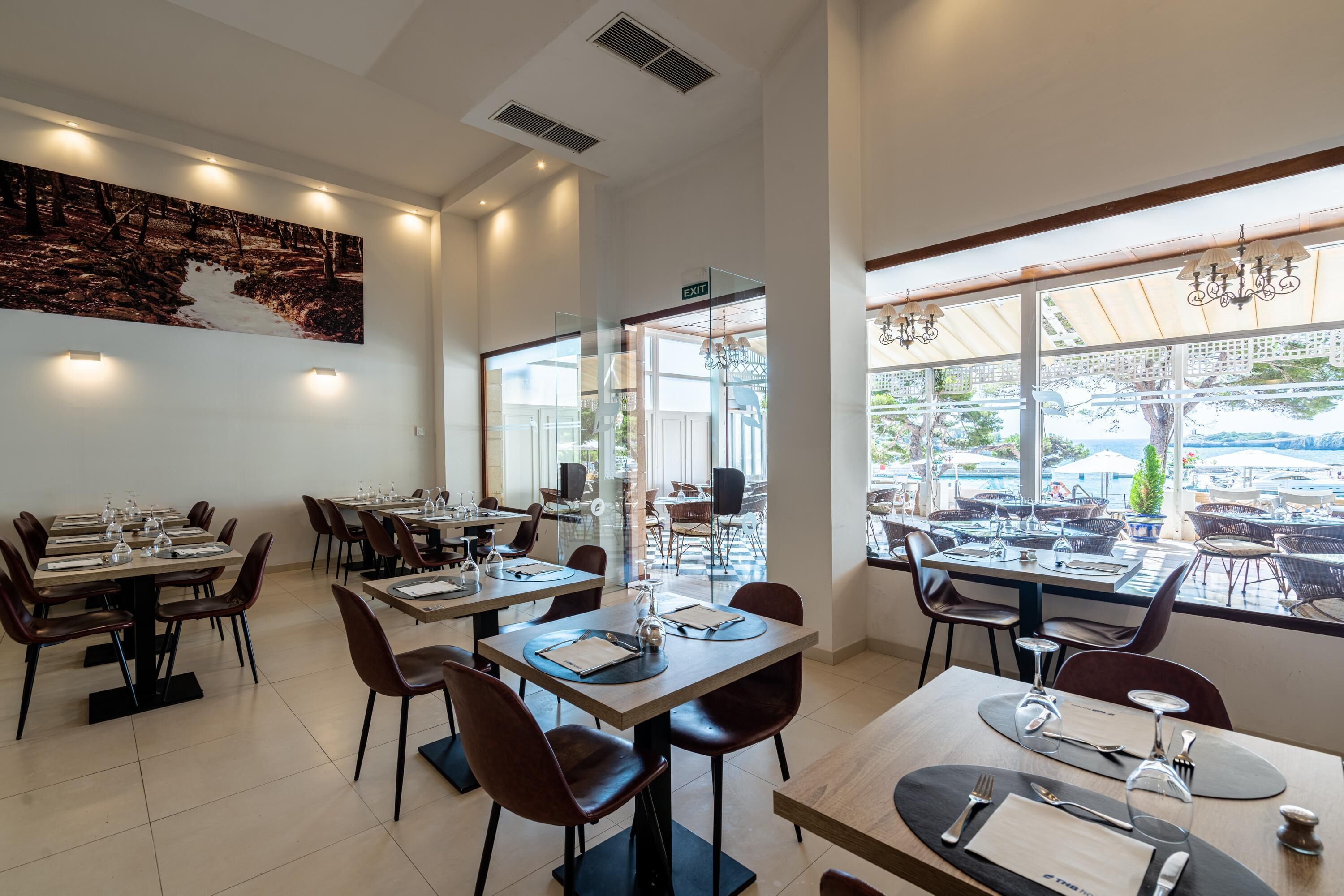 Ein Restaurant mit mehreren Tischen und Stühlen, einem großen Gemälde an der Wand und großen Fenstern mit Blick ins Freie.