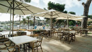 Café al aire libre con mesas y sillas bajo sombrillas grandes, con vista a un puerto deportivo lleno de barcos.