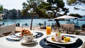 A table with breakfast items, including a sandwich, coffee, and fruit, overlooking a body of water with a view of trees and buildings. - Hotel THB Felip - Adults Only (Manacor)