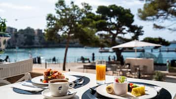 A table with breakfast items, including a sandwich, coffee, and fruit, overlooking a body of water with a view of trees and buildings.
